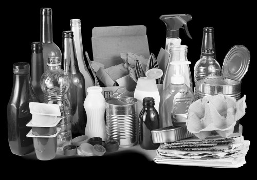 A collection of various discarded items positioned on a flat surface, including multiple glass bottles of different shapes and sizes, some with caps and others without, alongside a few plastic containers and bottles, some with spray nozzles. There are also several aluminum cans, paper and cardboard waste, including a crumpled paper bag and flattened cardboard sheets. The items appear to be gathered for disposal or recycling, with some objects stacked or leaning against each other. The background is dark, highlighting the reflective surfaces of the bottles and the varied textures of the waste materials. This arrangement of waste materials aligns with typical rubbish removal service tasks, such as those provided by Commercial Waste Removal Eltham, focused on clearing domestic or commercial waste in the Eltham area, reflecting a mix of recyclable and general rubbish often collected from local properties in southeast London.