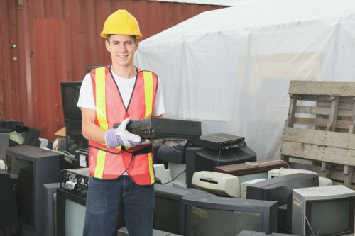 A young man wearing a yellow safety helmet, a high-visibility safety vest with pink and yellow reflective stripes over a white T-shirt, and work gloves, is standing outdoors near a collection of discarded electronic equipment including old television monitors, a printer, and other electronic waste. The background features a red wooden fence and a white fabric canopy or temporary shelter. The man is smiling and holding a black electronic device, possibly a laptop or similar item, indicating a focus on waste collection or disposal services. The surrounding environment suggests a waste removal or recycling yard, consistent with the services offered by Commercial Waste Removal Eltham in the context of rubbish and electronic waste disposal in the Eltham area. The atmosphere appears daylight, natural, and professional, highlighting proper health and safety practices related to waste handling and removal.