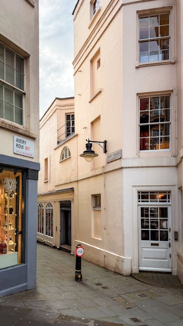 The image depicts a narrow, urban alleyway bordered by cream-colored, multi-story buildings with traditional sash windows and decorative architectural features, situated in a commercial area. In the f
