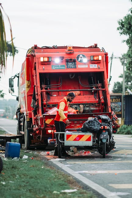 A municipal waste collection worker wearing a high-visibility yellow safety vest with reflective strips is seen emptying a black plastic garbage bag into the rear opening of a large red rubbish collec