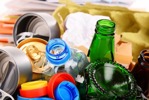 A close-up view of a collection of discarded recyclable bottles and containers, including green, blue, and clear glass and plastic bottles, some with lids and caps, alongside crumpled paper and cardboard packaging in the background. The items are scattered on a surface, likely outdoors or in a waste collection area, illustrating typical refuse that a professional rubbish removal service such as Commercial Waste Removal Eltham would handle. The containers have various textures and finishes, with some appearing smooth and glossy, reflecting light, while others are matte or crumpled. The scene captures the clutter associated with household or commercial waste awaiting collection, emphasizing the need for efficient rubbish removal in the local area. The background includes additional packaging and waste materials, all contributing to a realistic depiction of rubbish destined for disposal or recycling, consistent with the waste management services provided in Eltham and surrounding regions.